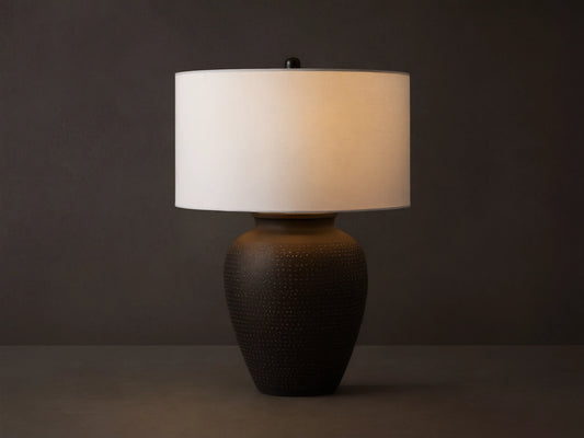 Howell Table Lamp with an embossed, dark powder-coated base and a wide, white cotton linen shade, on a wooden surface.