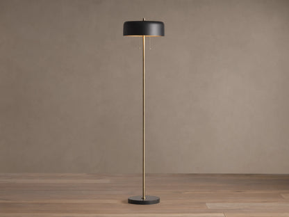 Oliver Floor Lamp: upright brass stem, black cylindrical shade with warm downward glow, black circular base on wooden floor.