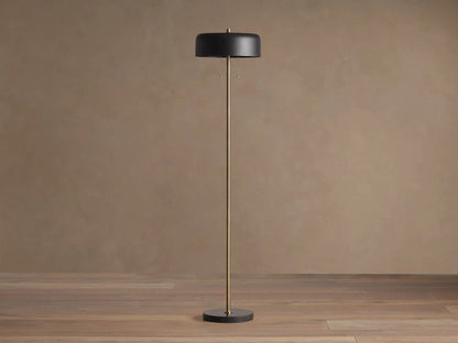Oliver Floor Lamp: upright brass stem, black cylindrical shade with warm downward glow, black circular base on wooden floor.