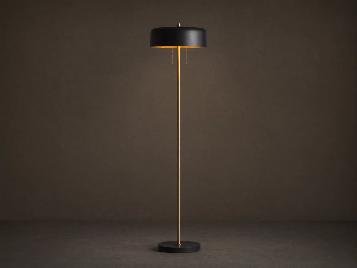 Oliver Floor Lamp: upright brass stem, black cylindrical shade with warm downward glow, black circular base on wooden floor.