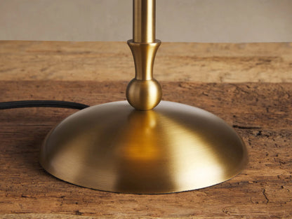 Archard Table Lamp with a brass finish, domed shade, slender stem, and round base, standing on a wooden table against a plain wall.