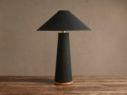 Ombra Table Lamp with black conical shade and tapering black fabric stem, brass accent, and teak wood base, on a wooden table.