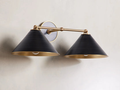 Arno Double Sconce with matte black cone-shaped shades, brass arm, polished brass interior, and round backplate on a wall.