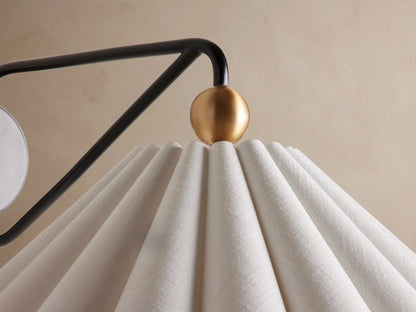 Haylen Sconce: Black iron wall light with antique brass accents, featuring a pleated, light-colored fabric cone shade, against a tan wall.