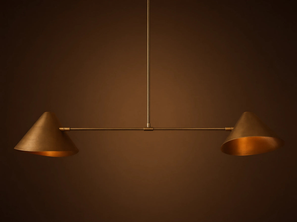 Simms Linear Chandelier with two downward-facing brass conical shades on a dark horizontal bar, suspended against a taupe wall.