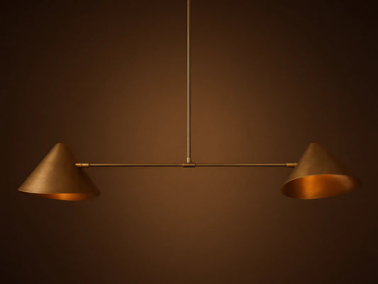 Simms Linear Chandelier with two downward-facing brass conical shades on a dark horizontal bar, suspended against a taupe wall.