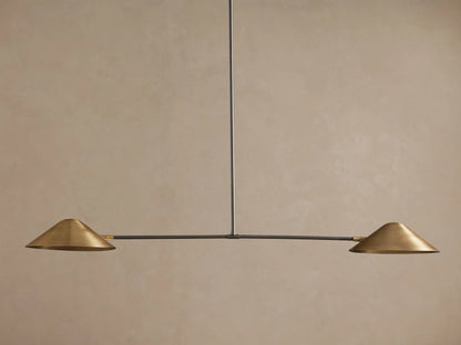 Simms Linear Chandelier with two downward-facing brass conical shades on a dark horizontal bar, suspended against a taupe wall.
