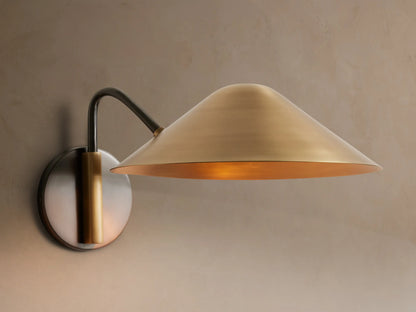 Simms Sconce: A large, conical polished brass shade emits a warm glow, attached to a dark curved arm & round backplate.