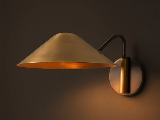 Simms Sconce: A large, conical polished brass shade emits a warm glow, attached to a dark curved arm & round backplate.