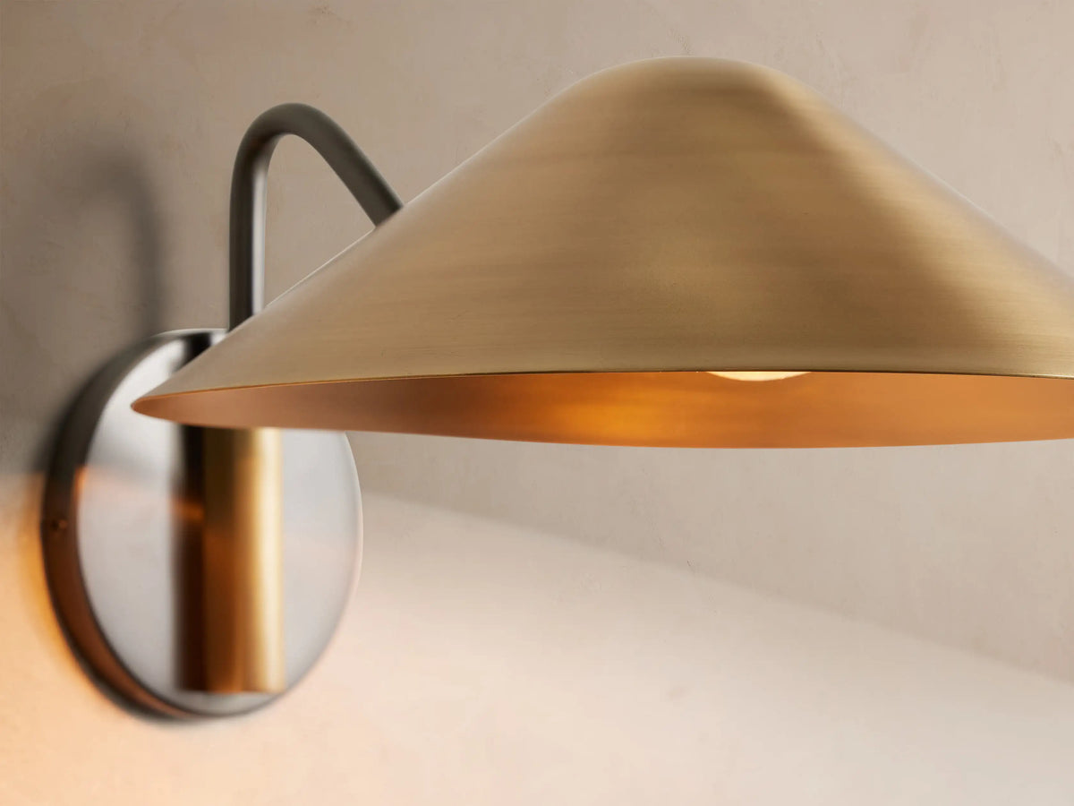 Simms Sconce, an antique brass wall lamp with a wide conical shade and dark curved arm, mounted on a round backplate.