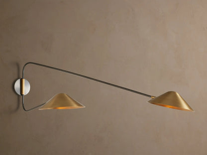 Simms Long Arm Double Sconce: Two antiqued brass conical lights with dark metal arms extending from a silver-toned backplate on a wall.
