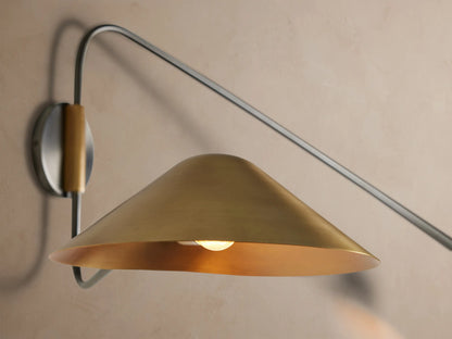 Simms Long Arm Double Sconce: dark curved metal structure with two polished brass conical shades against a beige wall.