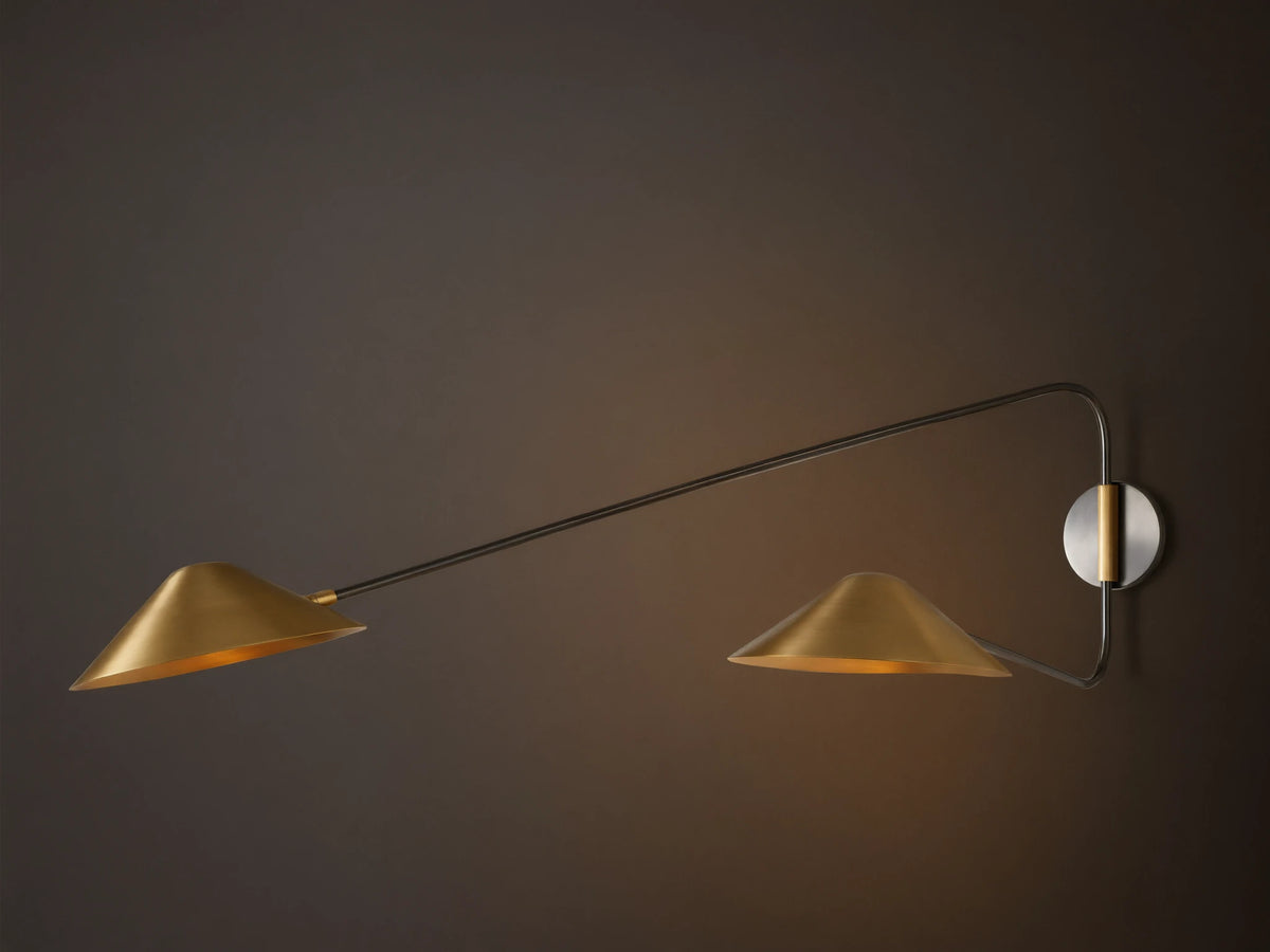 Simms Long Arm Double Sconce: Two antiqued brass conical lights with dark metal arms extending from a silver-toned backplate on a wall.