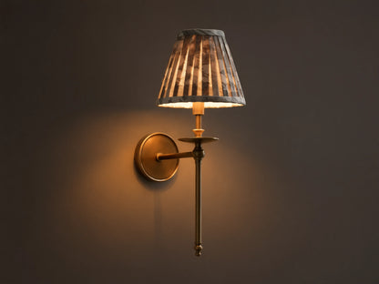 Front view of the lit antique brass Winslow Single Sconce, featuring a pleated dark patterned fabric shade, mounted on a wall.