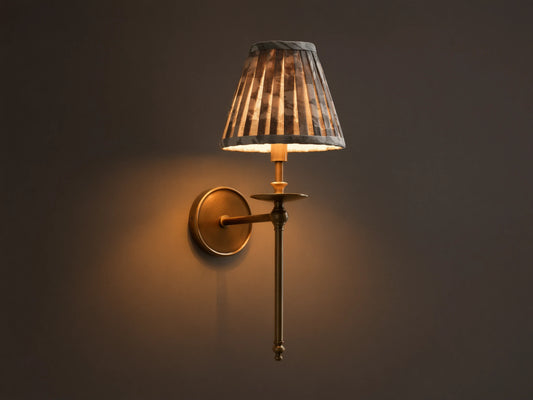 Front view of the lit antique brass Winslow Single Sconce, featuring a pleated dark patterned fabric shade, mounted on a wall.