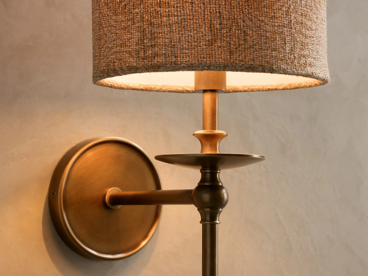 Front view of the Winslow Single Sconce with an antique brass finish and a brown fabric cylindrical shade, illuminated.