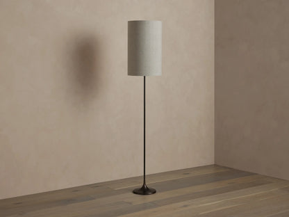 A tall, cylindrical floor lamp with a textured beige shade, emitting warm light. It has a slender black base, standing on a wooden floor.