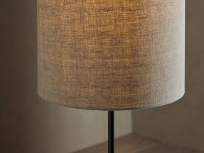 A tall, slender floor lamp with a cylindrical beige shade on a black metal stand, set against a light brown wall and wood floor.