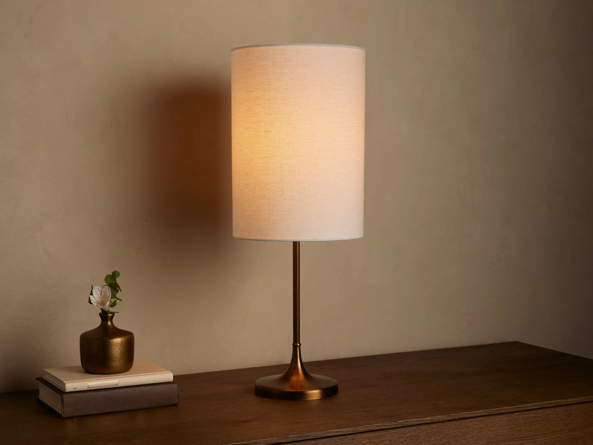Lit Lorelei Table Lamp with a flax linen cylindrical shade and antique brass base on a wooden dresser, casting a warm glow.