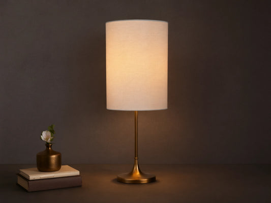 Lit Lorelei Table Lamp with a flax linen cylindrical shade and antique brass base on a wooden dresser, casting a warm glow.