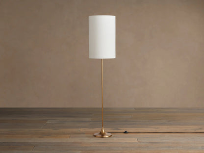 Lorelei Floor Lamp with antiqued brass finish, tall cylindrical white linen shade, standing on a wooden floor.