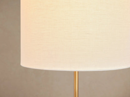 Antiqued brass Lorelei Floor Lamp, slender stem, round base, and tall white linen cylindrical shade on wood floor.