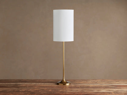 Lorelei Table Lamp with slender brass stem and round base, topped with an illuminated white cylindrical shade on a wooden table.