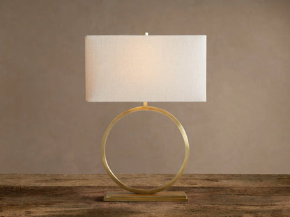 Rhys Table Lamp: Illuminated rectangular light fabric shade atop a gold circular hoop base on a rustic wood surface.
