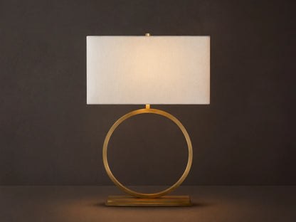 Rhys Table Lamp: Illuminated rectangular light fabric shade atop a gold circular hoop base on a rustic wood surface.