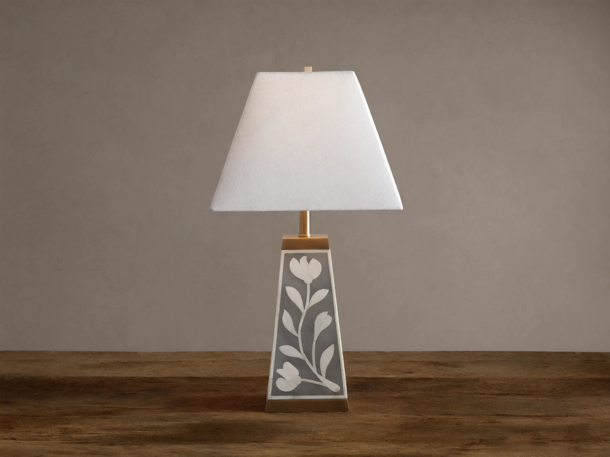 Divya Table Lamp with white fabric shade, gray base with white bone inlay floral motifs, brass trim, on wooden table.