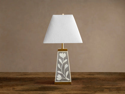 Divya Table Lamp with white fabric shade, gray base with white bone inlay floral motifs, brass trim, on wooden table.