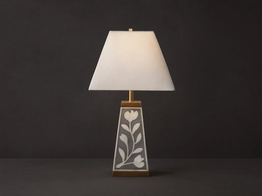 Divya Table Lamp with white fabric shade, gray base with white bone inlay floral motifs, brass trim, on wooden table.