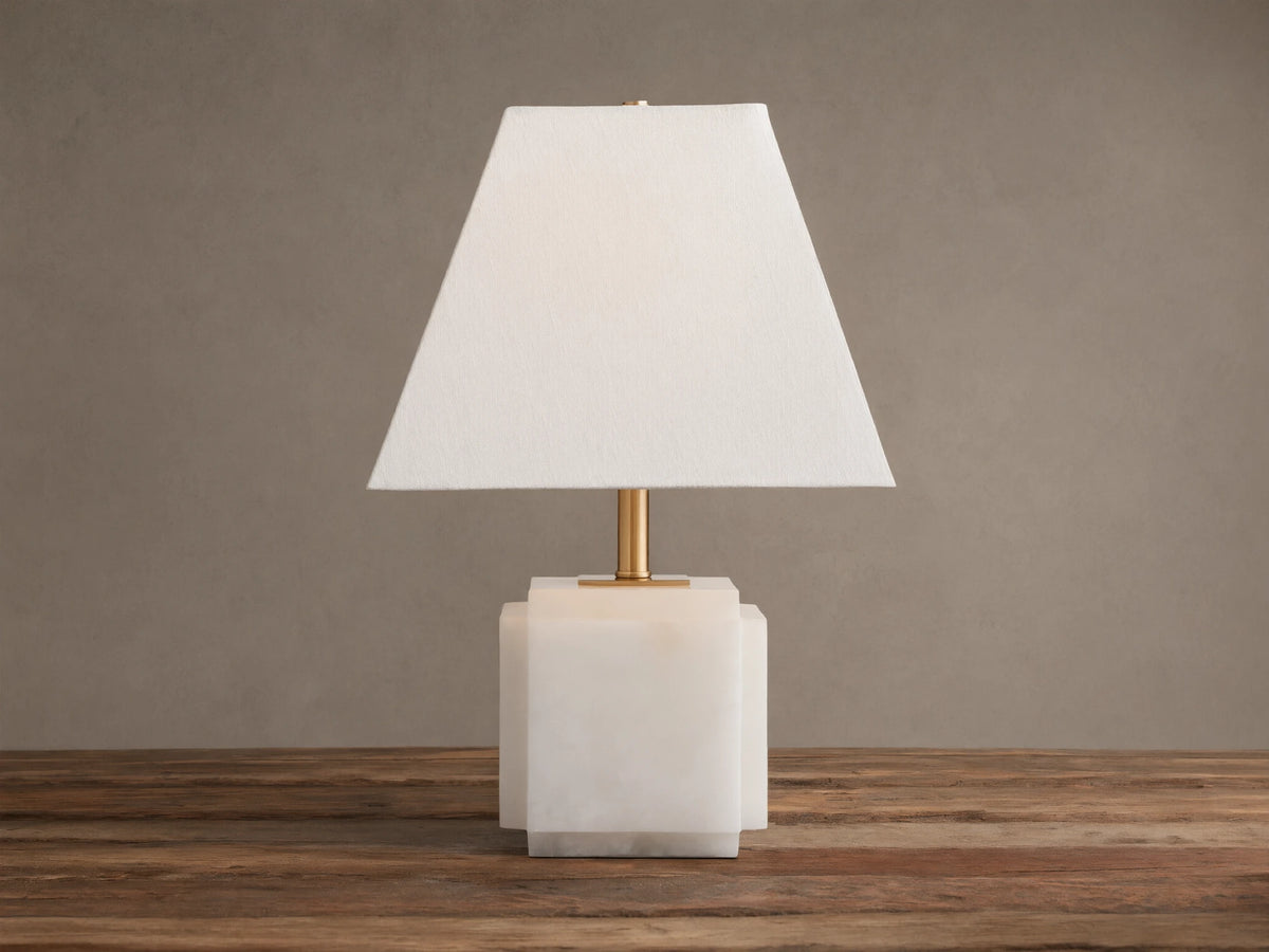 Fatima Table Lamp with a white natural stone base, gold stem, and tapered white linen shade, glowing softly on a wooden table.