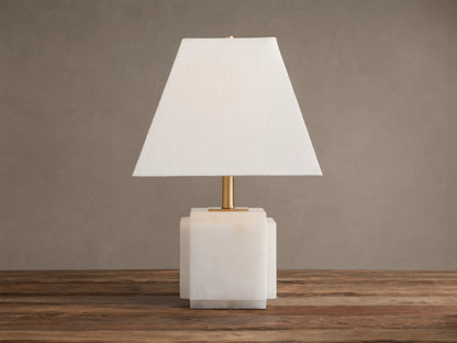 Fatima Table Lamp with a white natural stone base, gold stem, and tapered white linen shade, glowing softly on a wooden table.