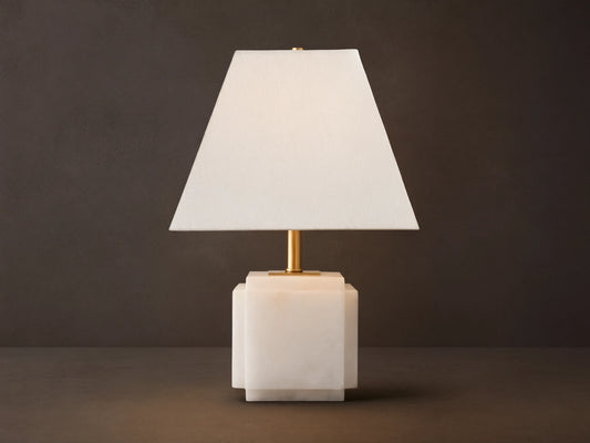 Fatima Table Lamp with a white natural stone base, gold stem, and tapered white linen shade, glowing softly on a wooden table.