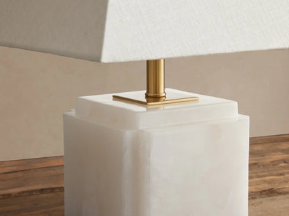 Fatima Table Lamp: White tapered linen shade, brass stem, and light natural stone block base on a rustic wooden table.