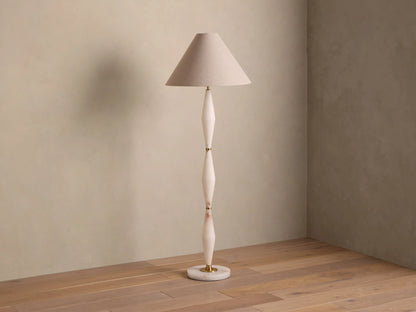 Full view of a floor lamp with a beige conical shade, off-white sculpted stem, and gold accents, casting a warm glow.