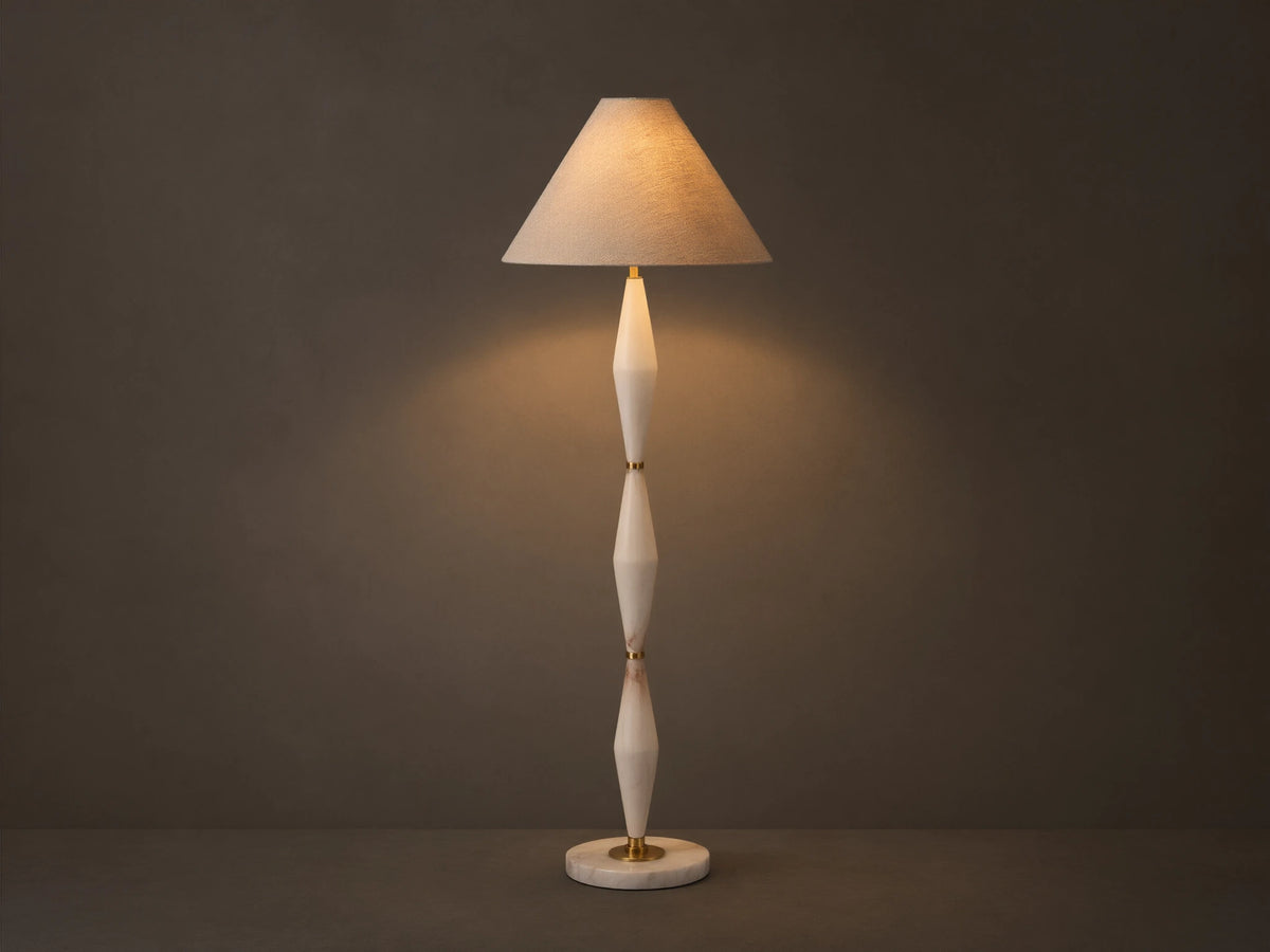 Full view of a floor lamp with a beige conical shade, off-white sculpted stem, and gold accents, casting a warm glow.