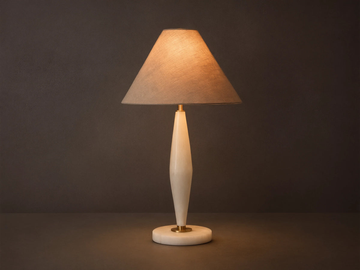 Front view of a lit table lamp with a conical beige fabric shade and a tapered creamy alabaster base with brass details.