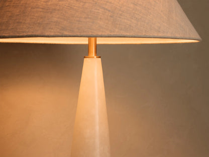 Close-up of a table lamp with a gray fabric lampshade and a gold-toned stem over a light-colored conical base, emitting warm light.