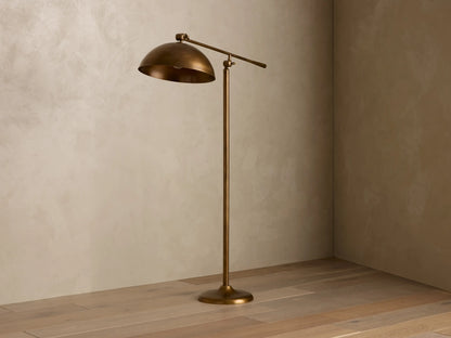 Interior view of a bronze floor lamp with a domed shade angled down, emitting a warm glow, standing on a wooden floor in a room corner.