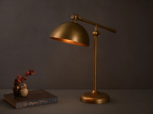 Front view of an illuminated brass industrial-style adjustable table lamp with a domed shade on a wooden surface.