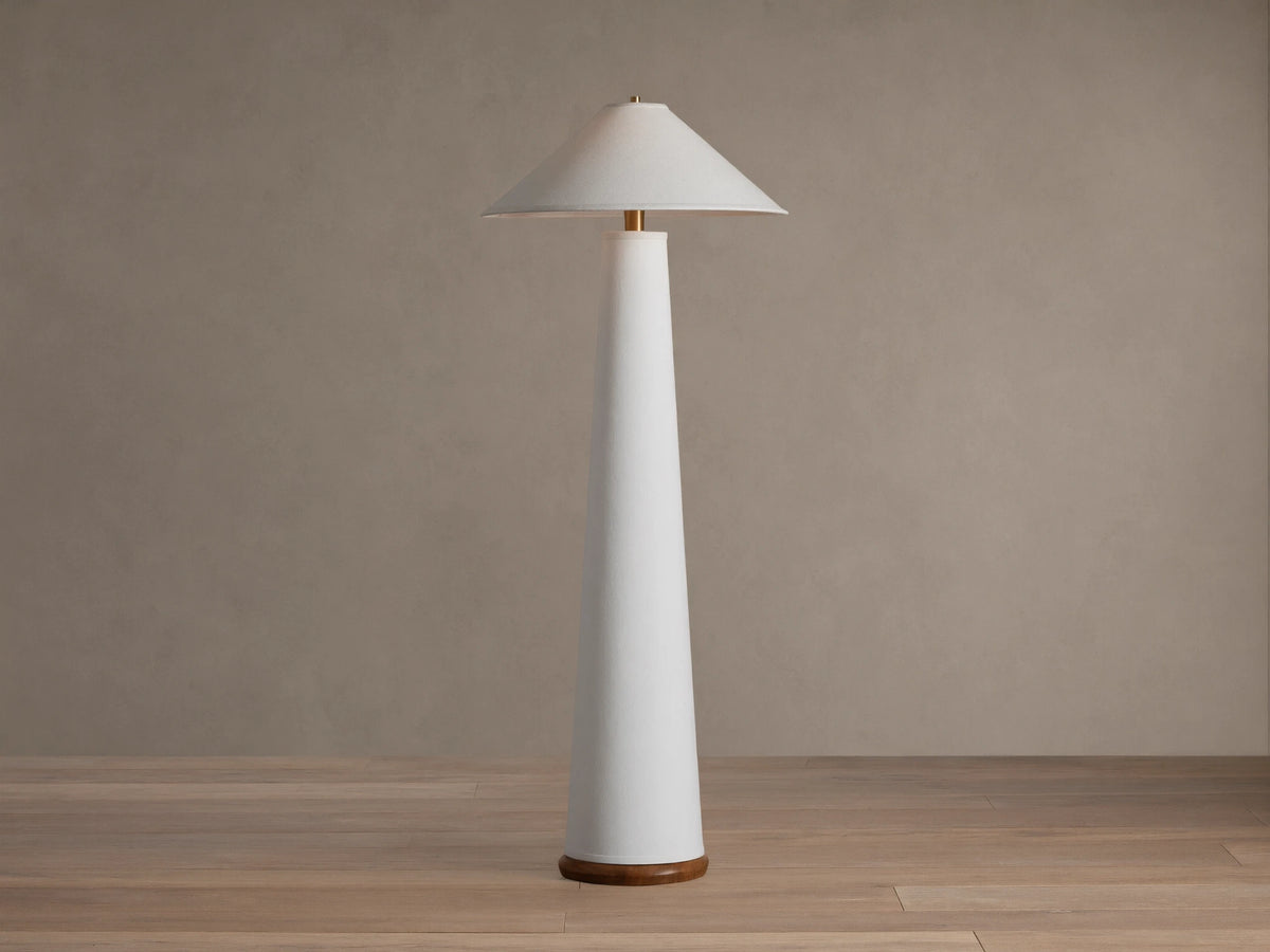 Ombra Floor Lamp: a tall white conical lamp with a white linen shade, teak base, and antiqued brass accents, on a wooden floor.