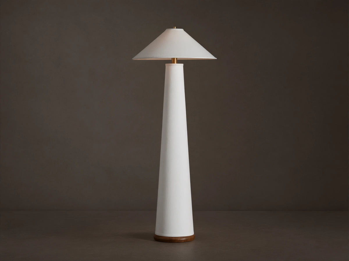 Ombra Floor Lamp: a tall white conical lamp with a white linen shade, teak base, and antiqued brass accents, on a wooden floor.