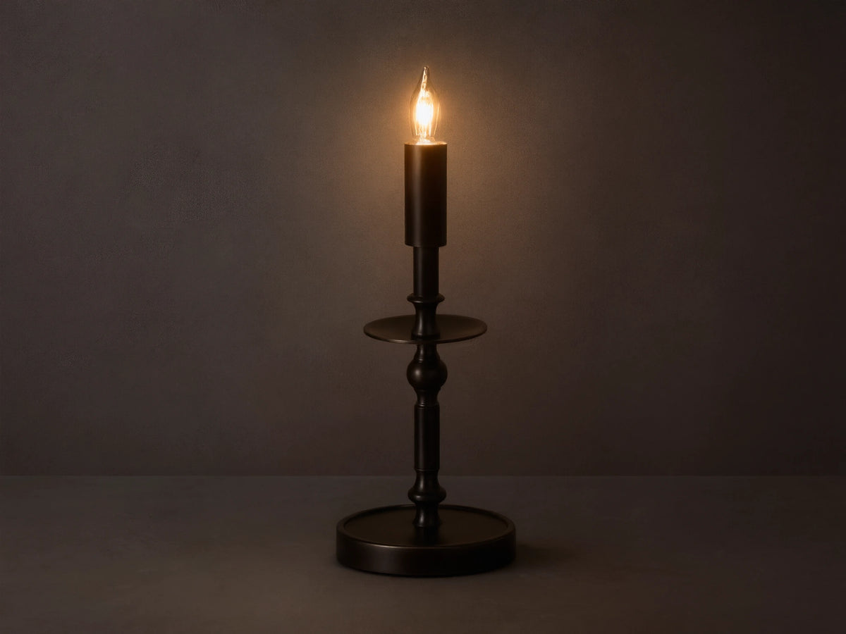Front view of the illuminated bronze Winslow Candlestick Table Lamp, on a wooden table with books and a small vase.