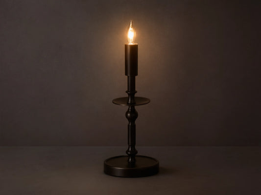 Front view of the illuminated bronze Winslow Candlestick Table Lamp, on a wooden table with books and a small vase.