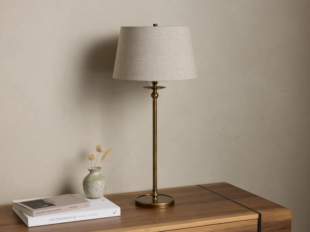 Front view of the Winslow Table Lamp with an antique brass base and light grey fabric shade, illuminated, on a wooden table.