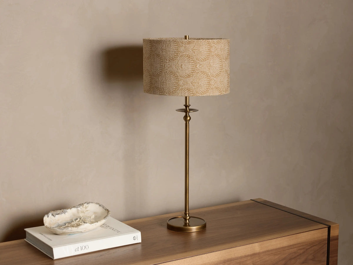 Close-up of the lit Winslow Table Lamp, highlighting its textured tan fabric shade and antique brass base with a warm glow.