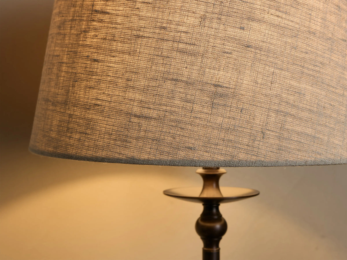 Front view of the Winslow Table Lamp, with a light natural fabric shade and dark bronze iron base, illuminated on a wooden table.