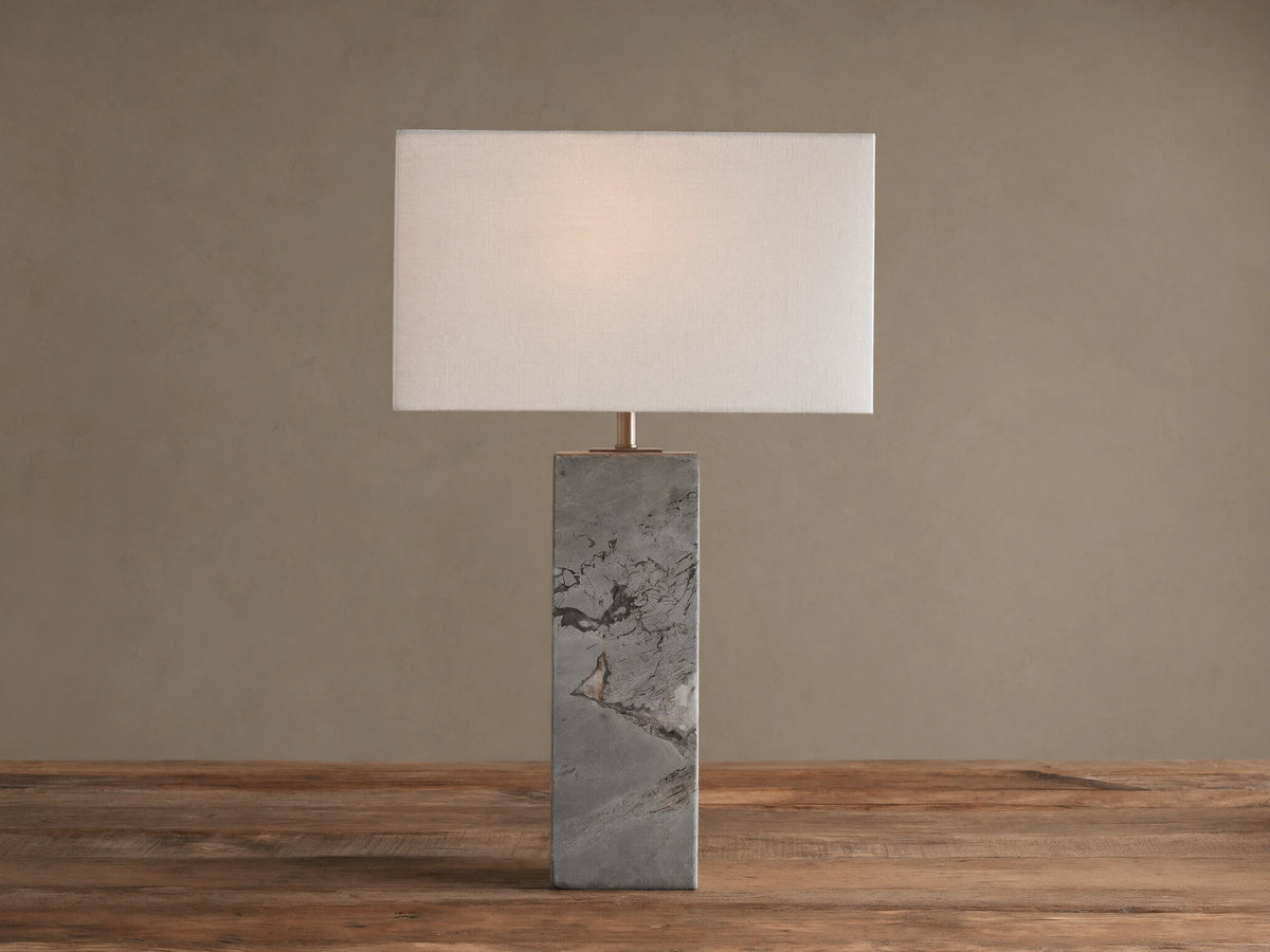 Oren Table Lamp: Rectangular white linen shade over a gray stone base with dark veining, sitting on a rustic wooden table.